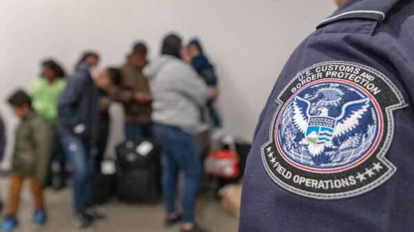 Badge on Customs and Border Patrol officer's uniform in focus with families lined up in background