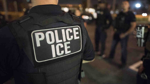 Photo is taken at night. The back of a black police vest is shown in the foreground with white letters reading "POLICE ICE"
