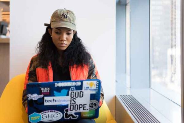 Individual in a green baseball cap sits in a yellow chair looking down at a laptop computer. The wall behind the person is white, with a grey window frame in the right third of the frame.