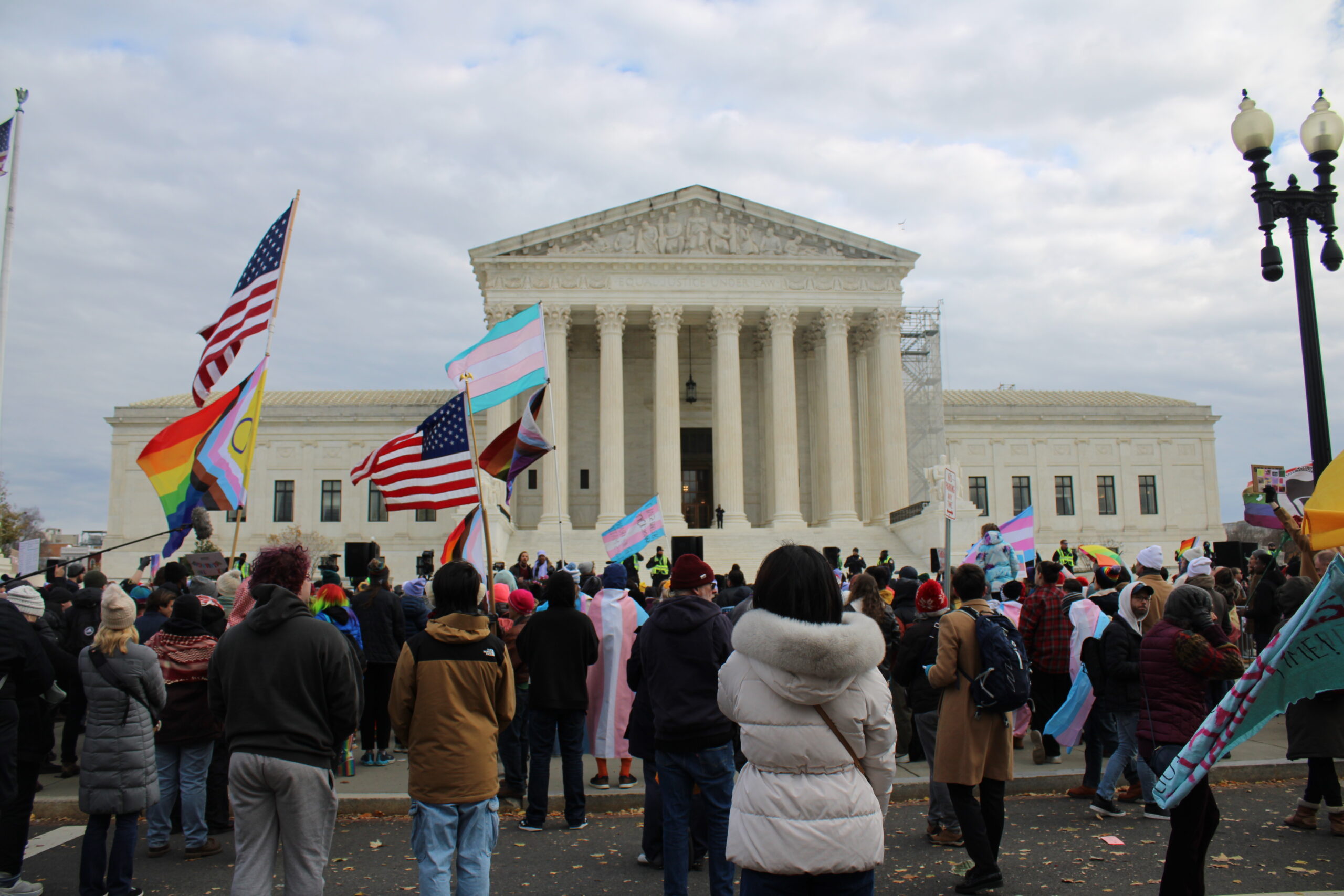 Protestors stand outside the Supreme Court of the United States with Trans flags and American flags waving above them.