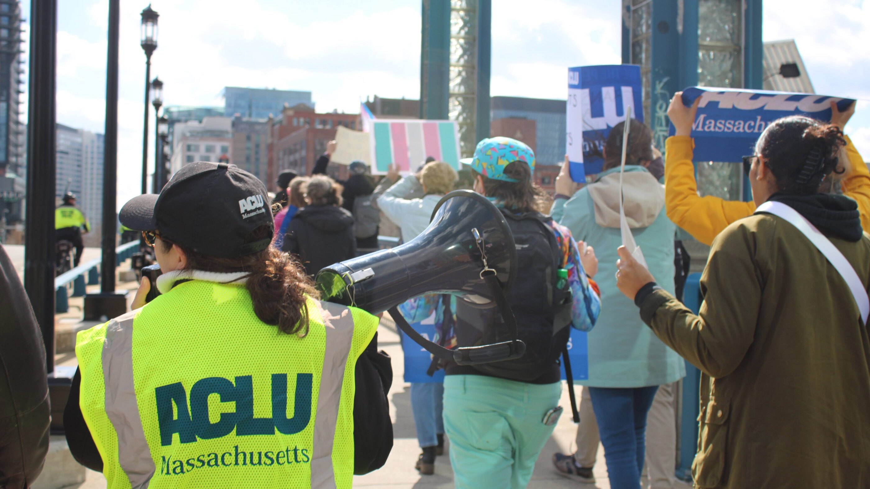 People marching in a protest; a woman wearing a yellow ACLU of Massachusetts vest is visible in the foreground.