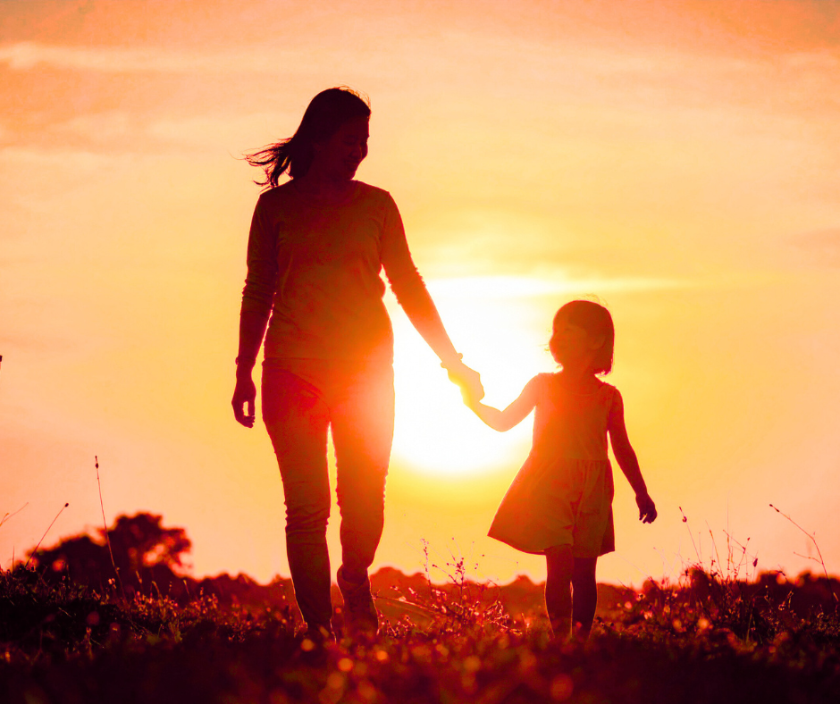 Mother and daughter hold hands walking toward camera with a sunset in the background.