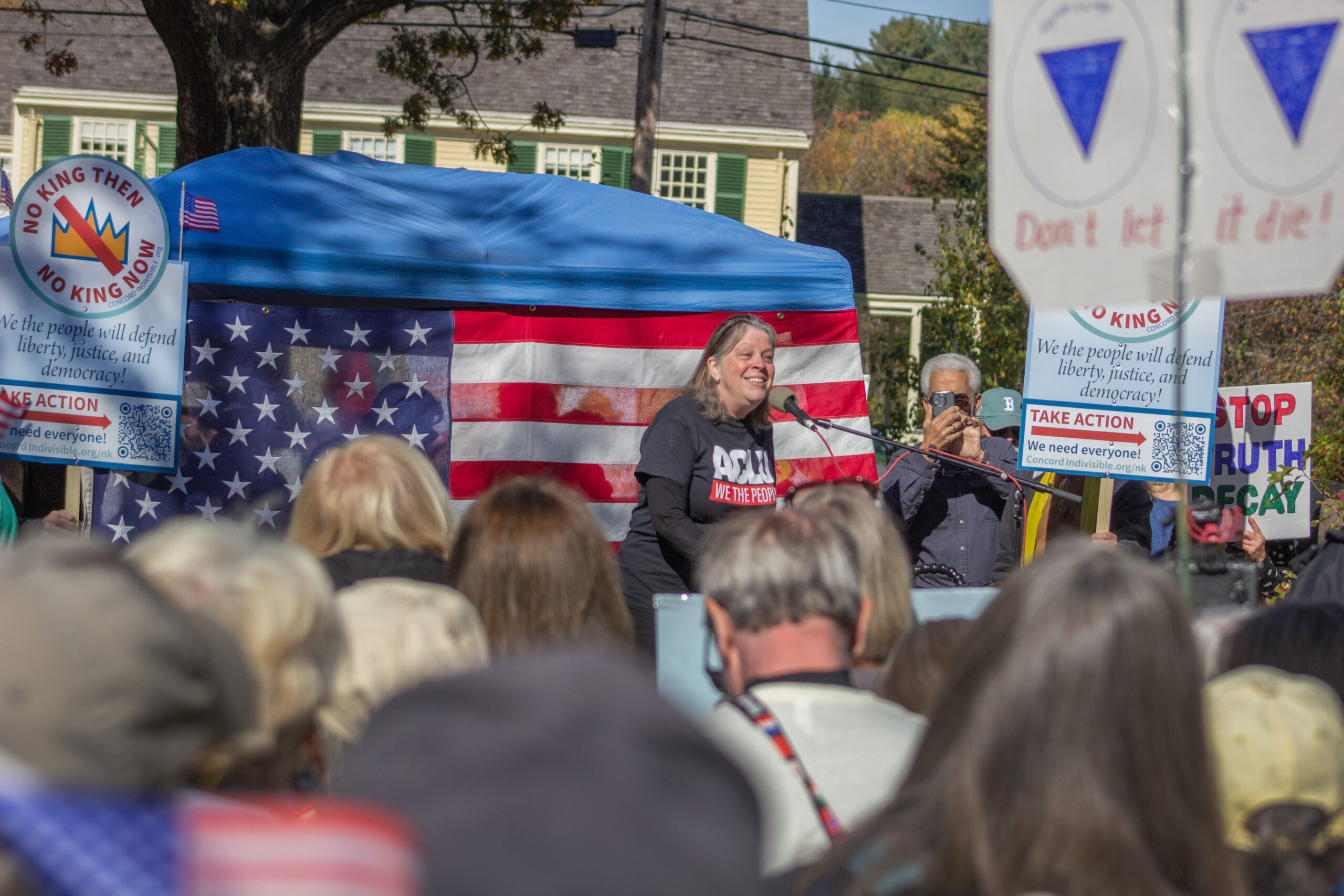 Carol Rose speaks to crowd in Concord, MA. An American Flag is hung up behind the speaker on stage with a blue tent over top.