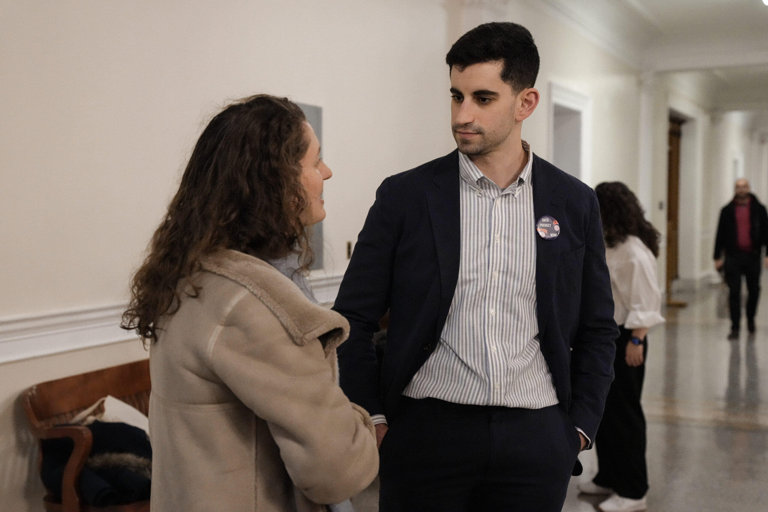 Two individuals are stood inside the Massachusetts Statehouse. The person on the left is turned to the side wearing a grey coat. On the right, a person is stood in a navy blue suit and light blue button up shirt with their head facing to the left listening.
