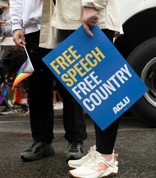 A protest sign reading "Free Speech, Free Country" is held by an individual in a white top and black pants.