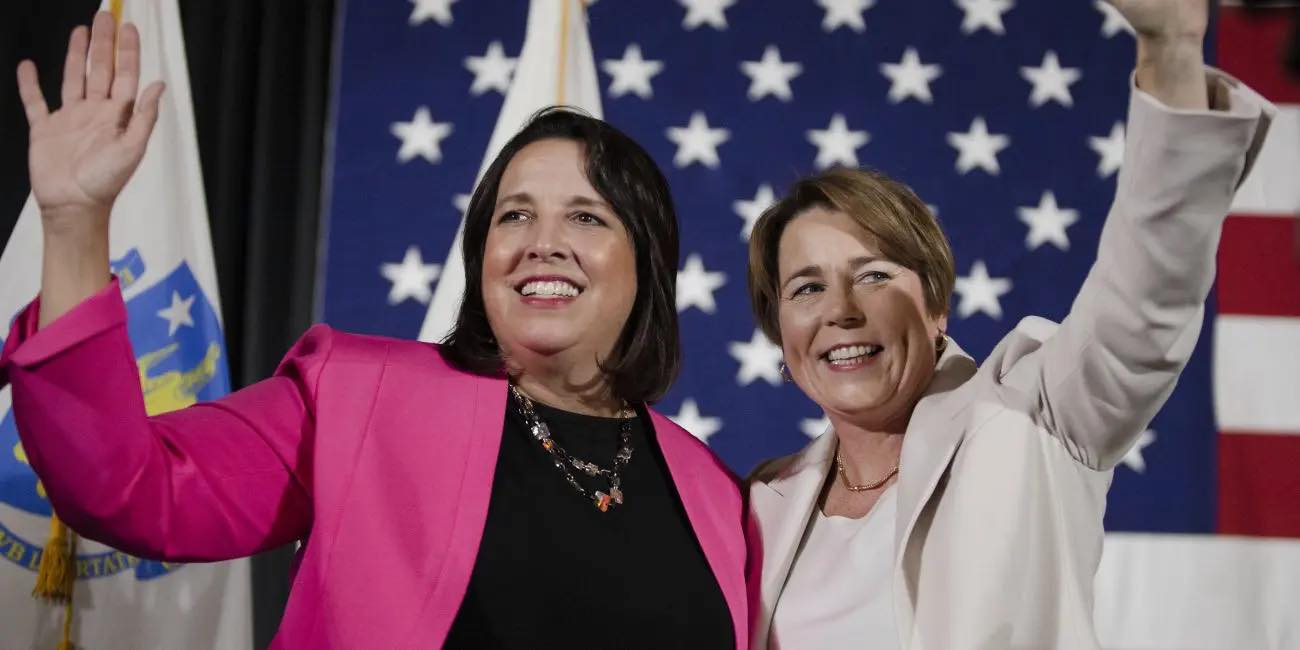 Governor Maura Healey and Lieutenant Governor Kim Driscoll stand side by side with their outer arms raised. They stand in front on an American Flag. Photo credit to Libby O'Neill