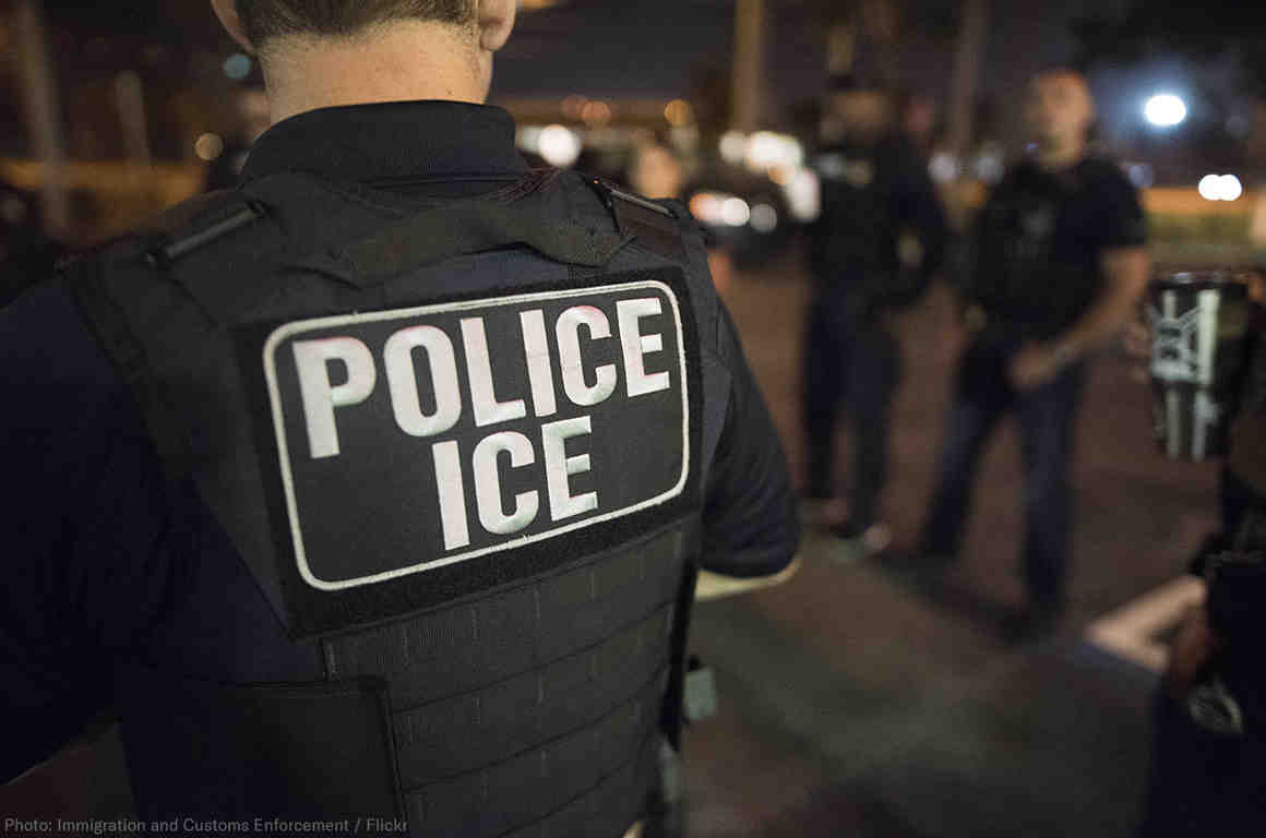 Photo is taken at night. The back of a black police vest is shown in the foreground with white letters reading "POLICE ICE" 