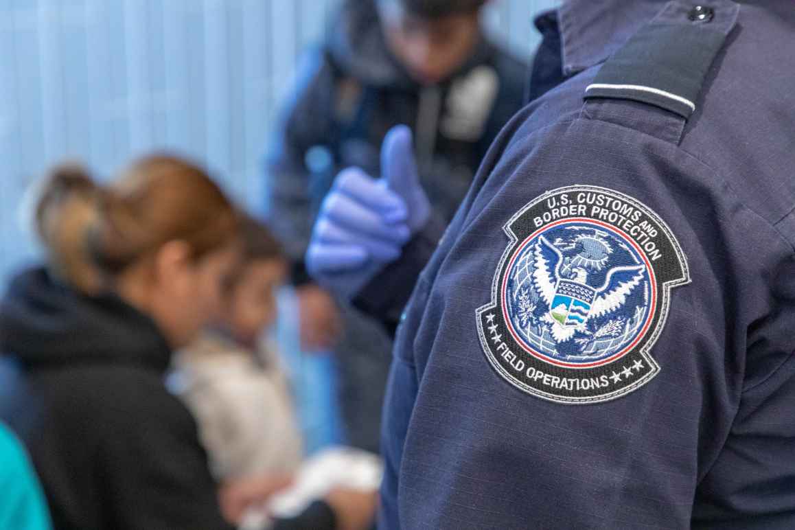 Badge on Customs and Border Patrol officer's uniform in focus with family being inspected in background