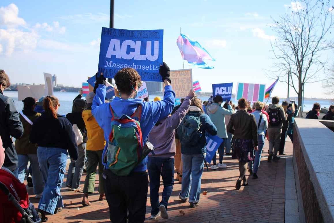 Group on individuals walking forward holding trans rights passport signs. A centered sign reads "ACLU of Massachusetts Hands Off Our Passports"