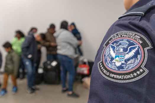 Badge on Customs and Border Patrol officer's uniform in focus with families lined up in background